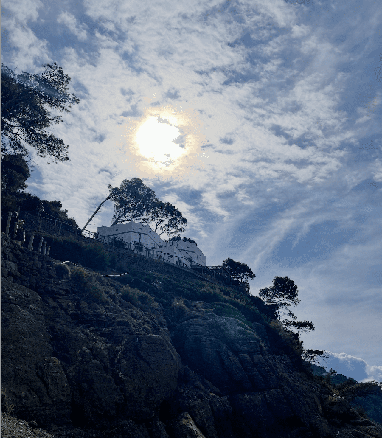 Image of a blue sky with wispy clouds and the sun blazing through and a steep mountain with palm trees silhouetted against the light